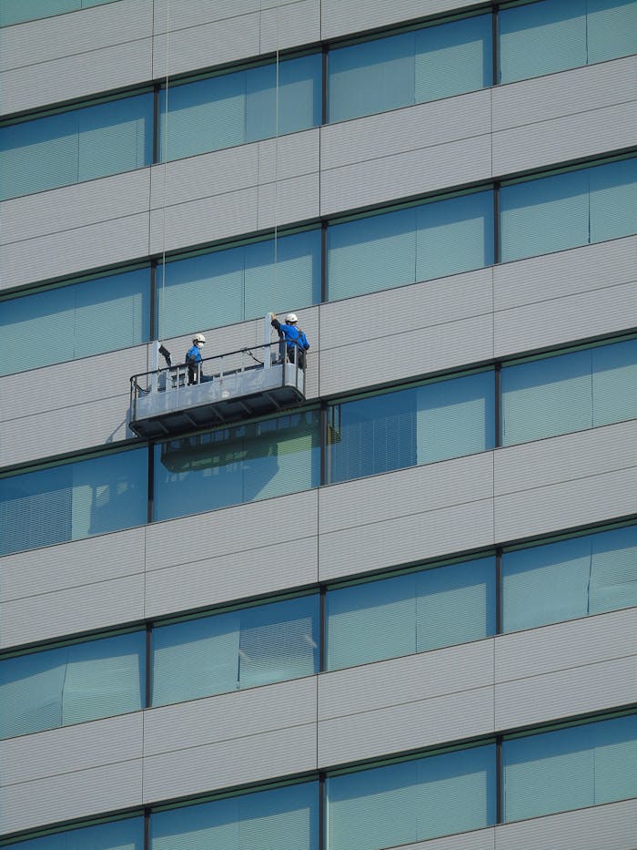 services-02 Two workers clean windows on a modern glass building facade using a suspended platform.